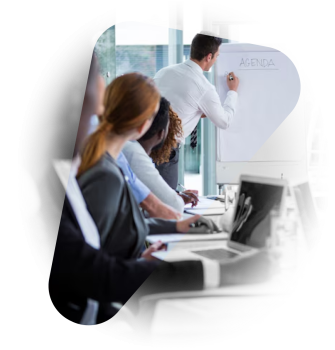 Na zdjęciu widzimy nowoczesne spotkanie biznesowe lub sesję szkoleniową w jasnym, przestronnym biurze. Kilka osób w eleganckich strojach siedzi przy stole konferencyjnym, skupiając uwagę na mężczyźnie stojącym przy flipcharcie. Na tablicy widnieje słowo „AGENDA”, co sugeruje rozpoczęcie prezentacji lub omówienie planu działania. Uczestnicy mają przed sobą laptopy, a przez duże okna wpada naturalne światło.